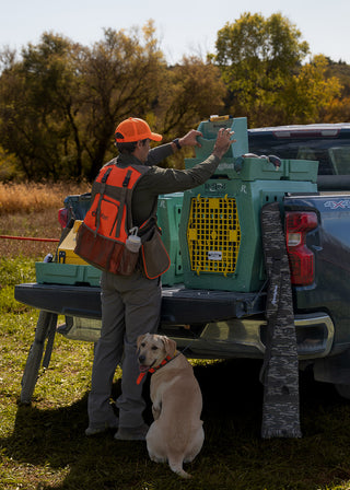 A man outdoors with his dog and his Limited Edition RuffLand x Remington Kennel collection. There is a Large RuffLand kennel, an Intermediate RuffLand kennel, a RuffLand Easy Rider Top Tray, and a RuffLand Water Hole. All in our signature colors: Go Green and Sunflower Yellow.