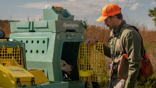 A man in outdoor hunting attire stands beside a RuffLand x Remington Dog Kennel  set up in a grassy field. The RuffLand Kennel is Go Green with Sunflower Yellow handles and doors with a Top Tray and Water Hole on top. A dog is visible inside the kennel.